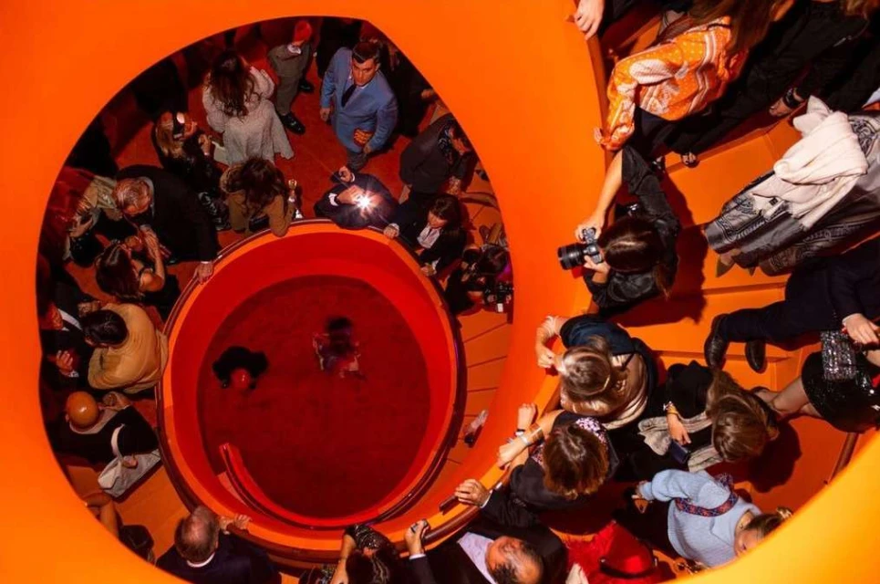 Overhead shot capturing a group of people on an orange spiral staircase, with some taking photos and others looking down the stairs.