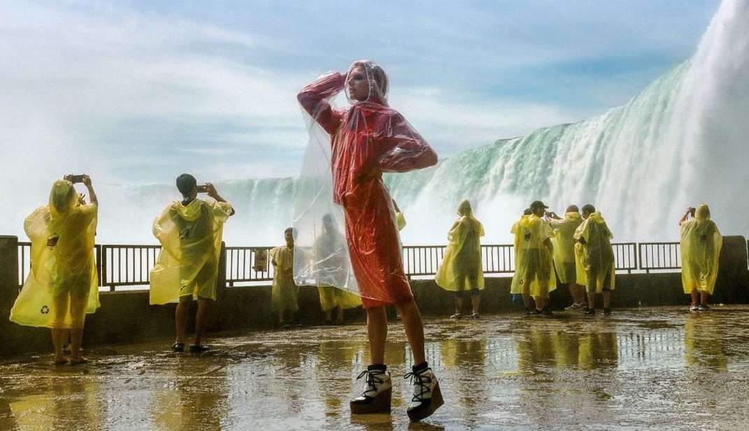 Fashion model poses in a red dress and clear rain poncho with a waterfall background as tourists in yellow ponchos take pictures.