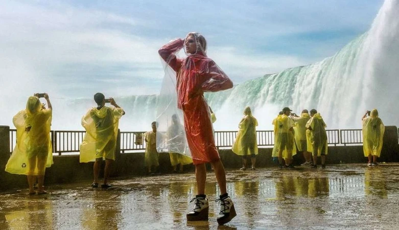 Fashion model poses in a red dress and clear rain poncho with a waterfall background as tourists in yellow ponchos take pictures.