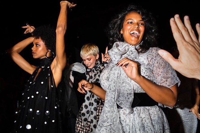 Three women in stylish dresses dance in a dimly lit setting with a raised hand visible. One wears polka dots, another a star pattern.