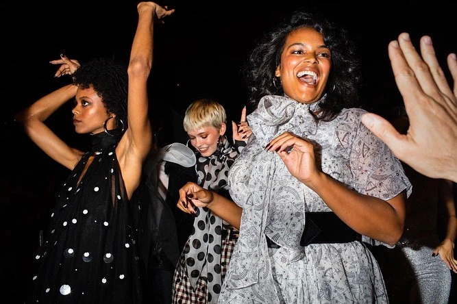 Three women in stylish dresses dance in a dimly lit setting with a raised hand visible. One wears polka dots, another a star pattern.