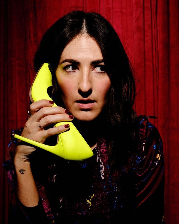 A woman with dark hair poses indoors, holding a yellow high-heel shoe to her ear like a phone, against a red curtain backdrop.