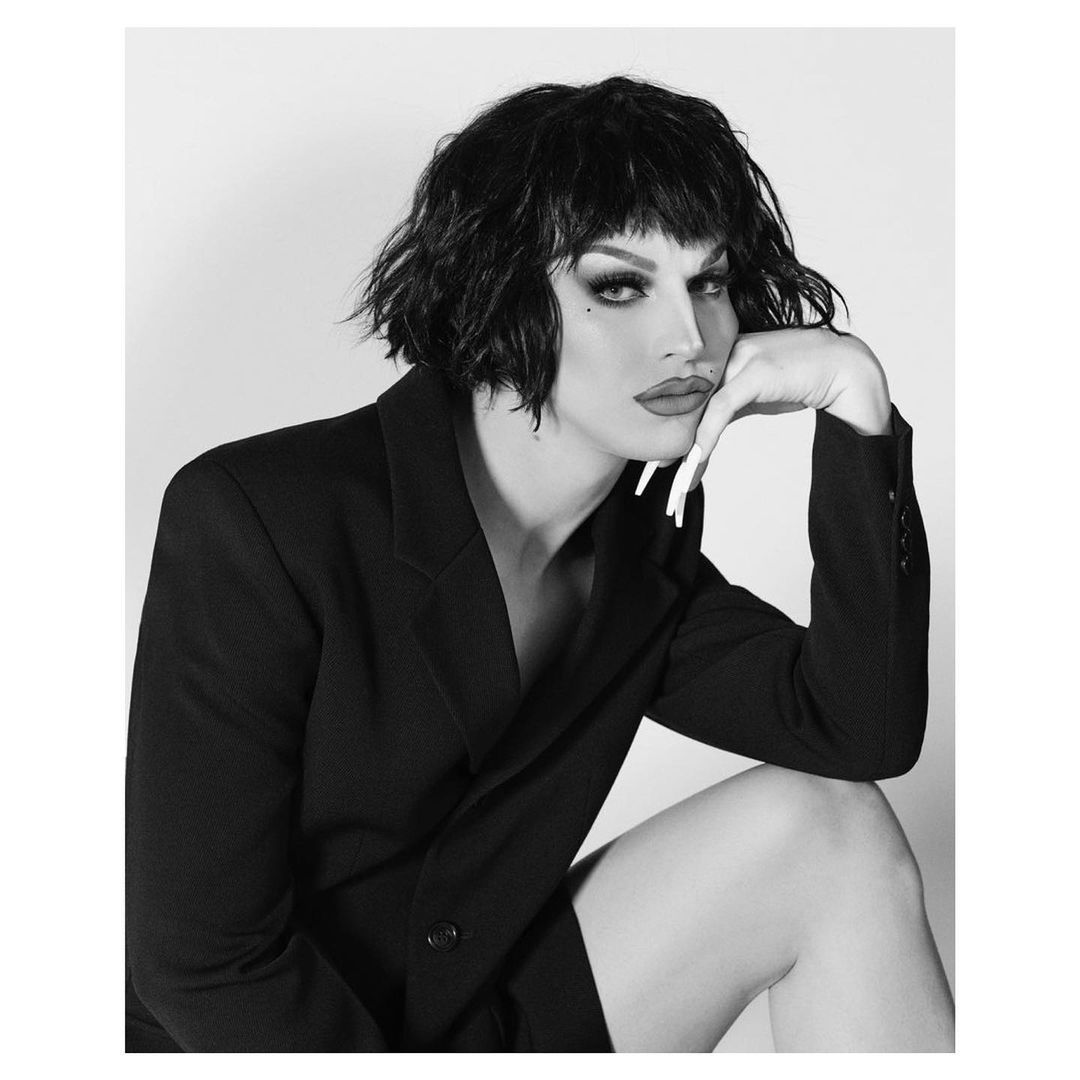 Black and white portrait of a person with dark hair in a black blazer. Studio shot, posed with hand on chin. Drag queen aesthetic.