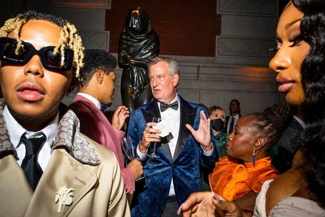 A group of well-dressed people gathers for an event in front of an art sculpture, creating a lively and fashionable atmosphere.