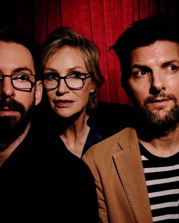 Three people pose indoors against a red curtain. Two bearded men and a woman in glasses and a tan blazer are posing for a photo.