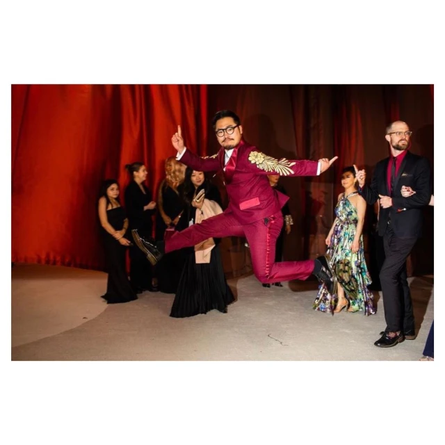 A man in a maroon suit is mid-air in a leaping pose, one leg tucked and the other extended, in front of a group of onlookers and red curtains.