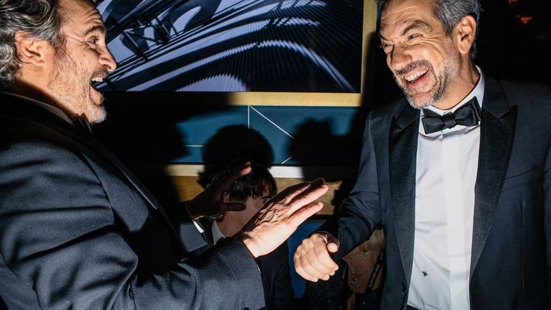 Two adult men in suits smiling and laughing at an event indoors. They are dressed formally with bow ties, and there is a digital art display behind them.