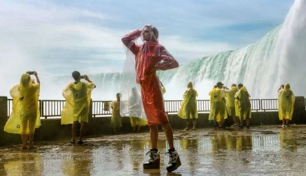 Model in translucent raincoat standing before a massive waterfall while tourists in yellow ponchos look on
