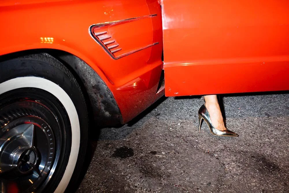 Close-up of a person wearing gold stiletto high heels stepping out of a bright red vintage car at night.