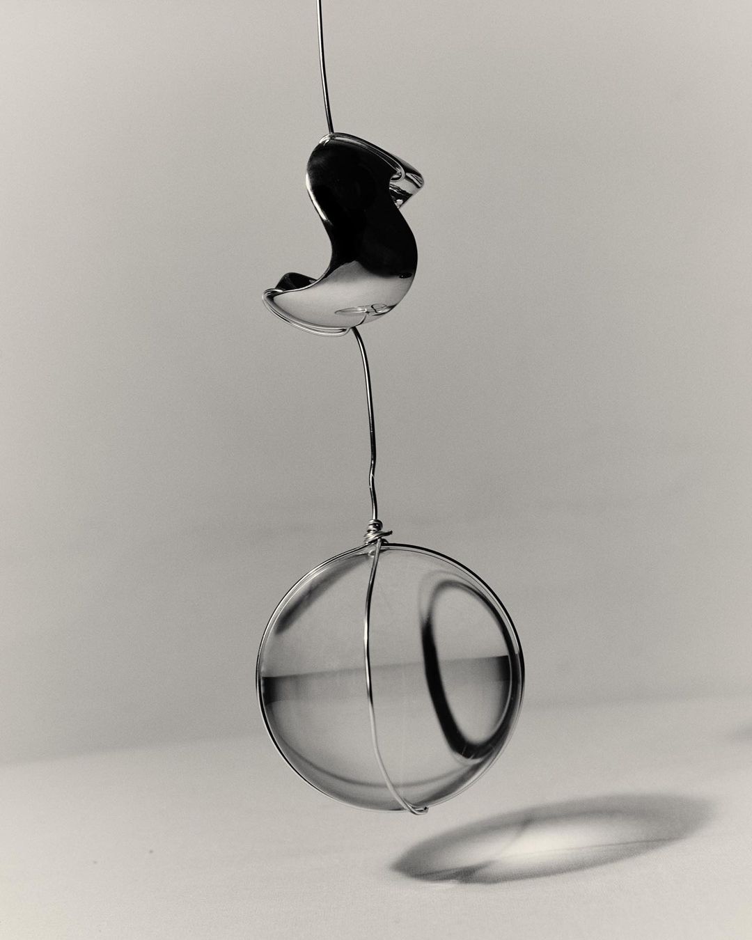 Still life of a clear glass sphere hanging from a thin wire against a gray gradient background with diffused lighting.