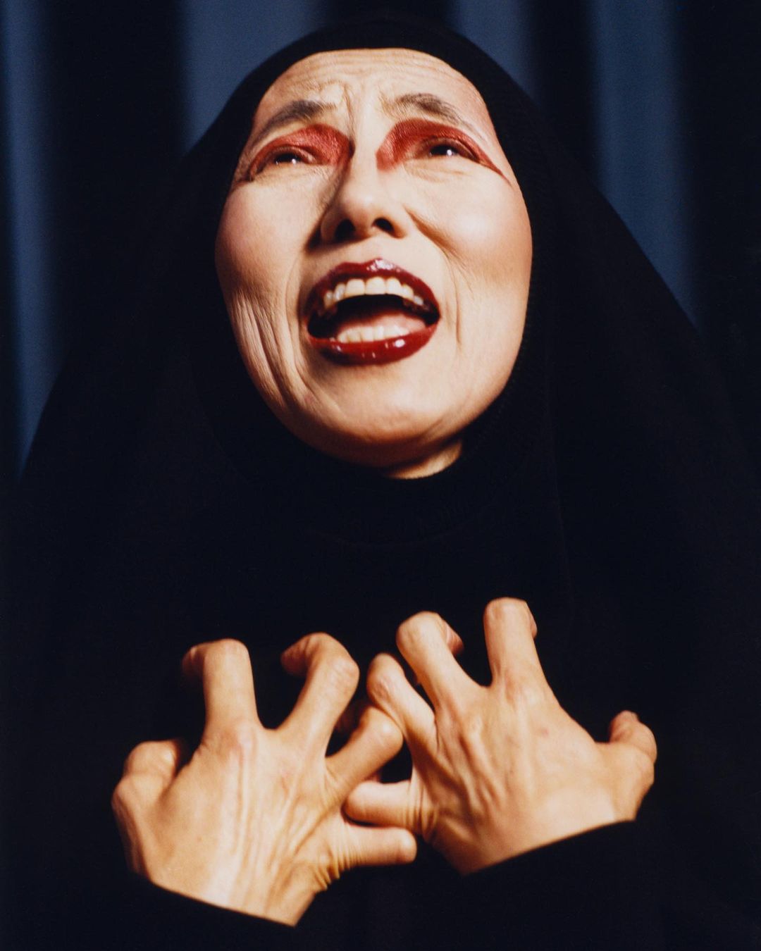 Dramatic portrait of a mature woman with exaggerated makeup, dressed in black, expressing a strong emotion with raised hands indoors.