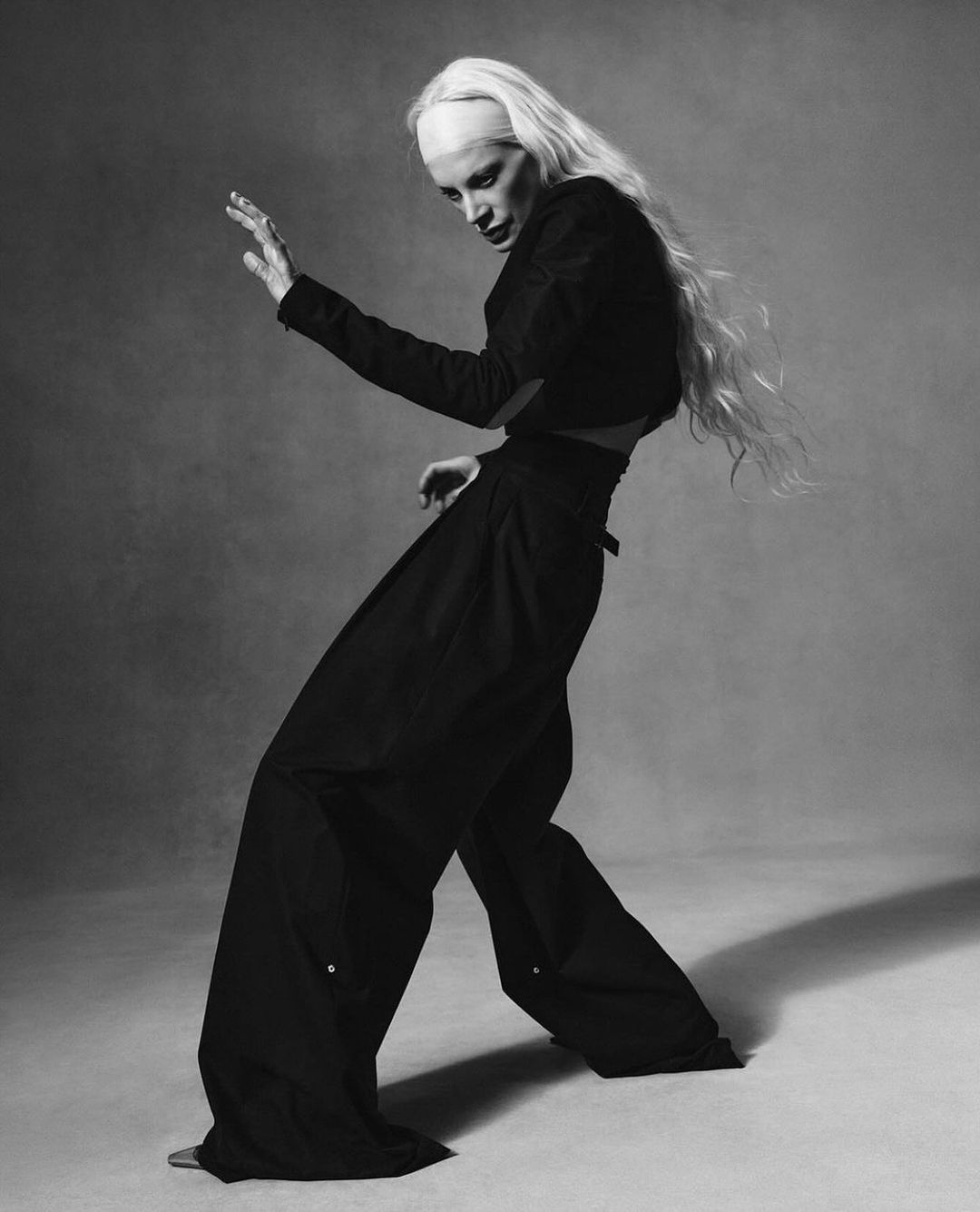 Monochrome fashion shot of a slender adult in dark clothing posing with outstretched arms, a bald head, and long white hair.