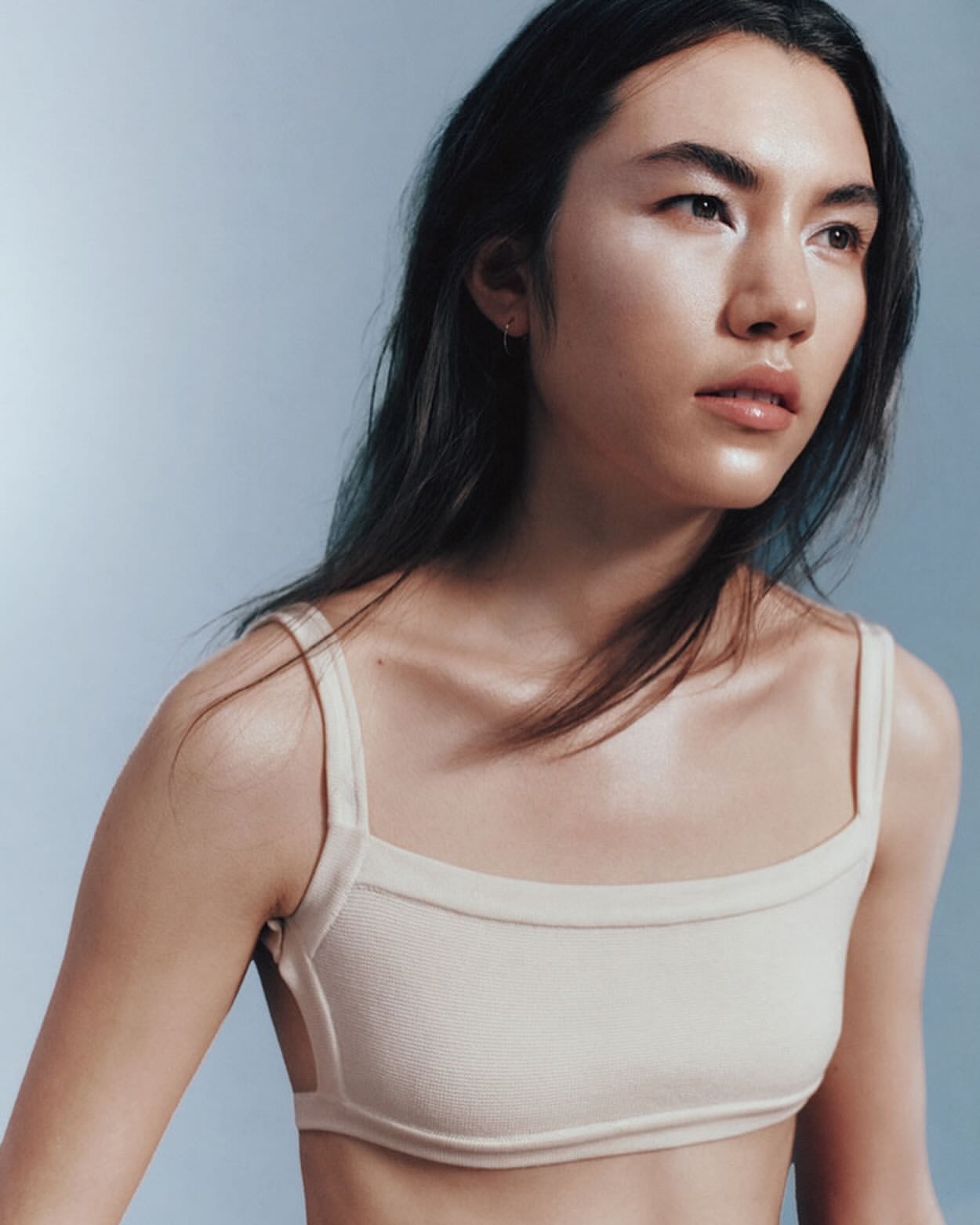 Portrait of an Asian woman wearing a beige knit bra top against a pale blue studio backdrop, with soft lighting and a medium shot.