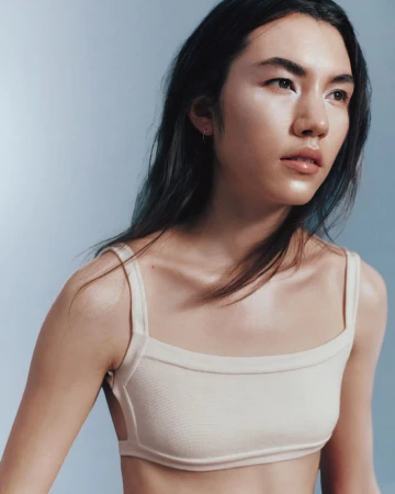 Portrait of an Asian woman wearing a beige knit bra top against a pale blue studio backdrop, with soft lighting and a medium shot.