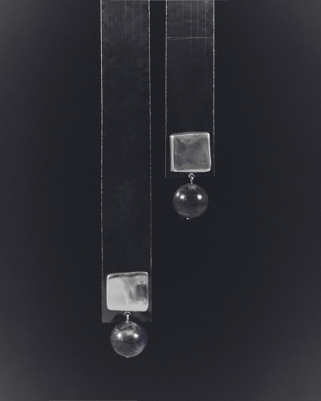 Abstract still life photograph of two geometric hanging objects. The composition features square and spherical forms against a dark backdrop.
