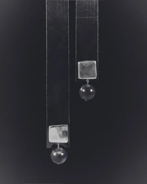 Abstract still life photograph of two geometric hanging objects. The composition features square and spherical forms against a dark backdrop.