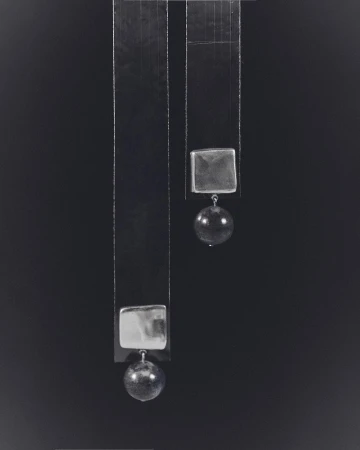 Abstract still life photograph of two geometric hanging objects. The composition features square and spherical forms against a dark backdrop.