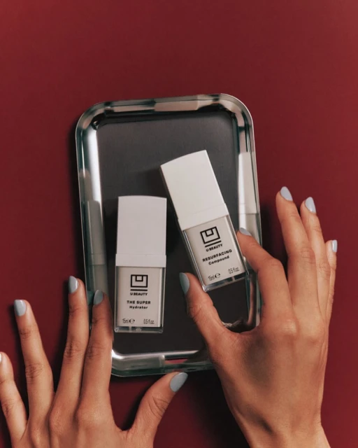 Two U Beauty skincare products are presented on a reflective silver tray against a red background, held by hands with light blue nail polish.
