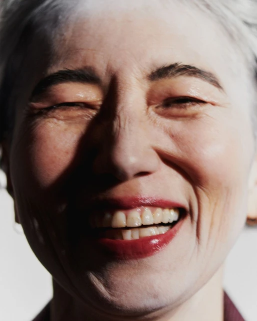 Close-up of a smiling elderly woman with white hair and red lipstick against a white background in a brightly lit studio environment.