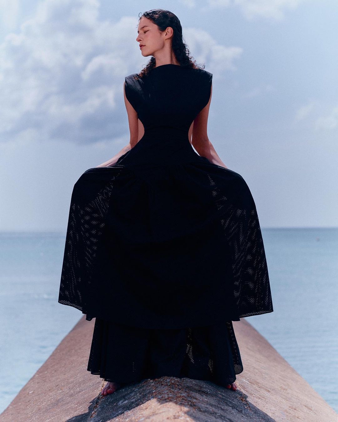 Woman in flowing black dress stands on a concrete pier under a cloudy sky, facing away from the camera with a serene ocean backdrop.