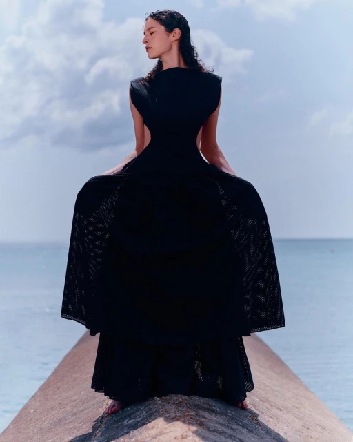 Woman in flowing black dress stands on a concrete pier under a cloudy sky, facing away from the camera with a serene ocean backdrop.