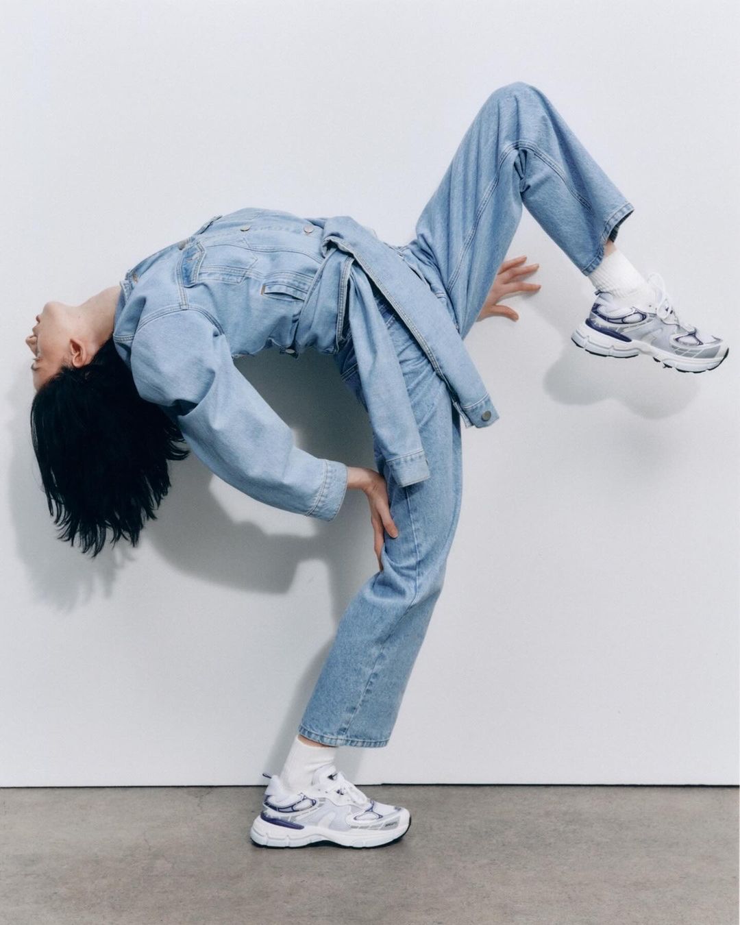 Woman in blue denim jumpsuit arches backward against a white wall, wearing white sneakers and socks, balancing on one foot.