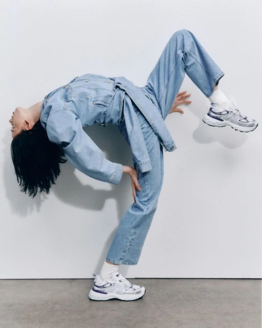 Woman in blue denim jumpsuit arches backward against a white wall, wearing white sneakers and socks, balancing on one foot.