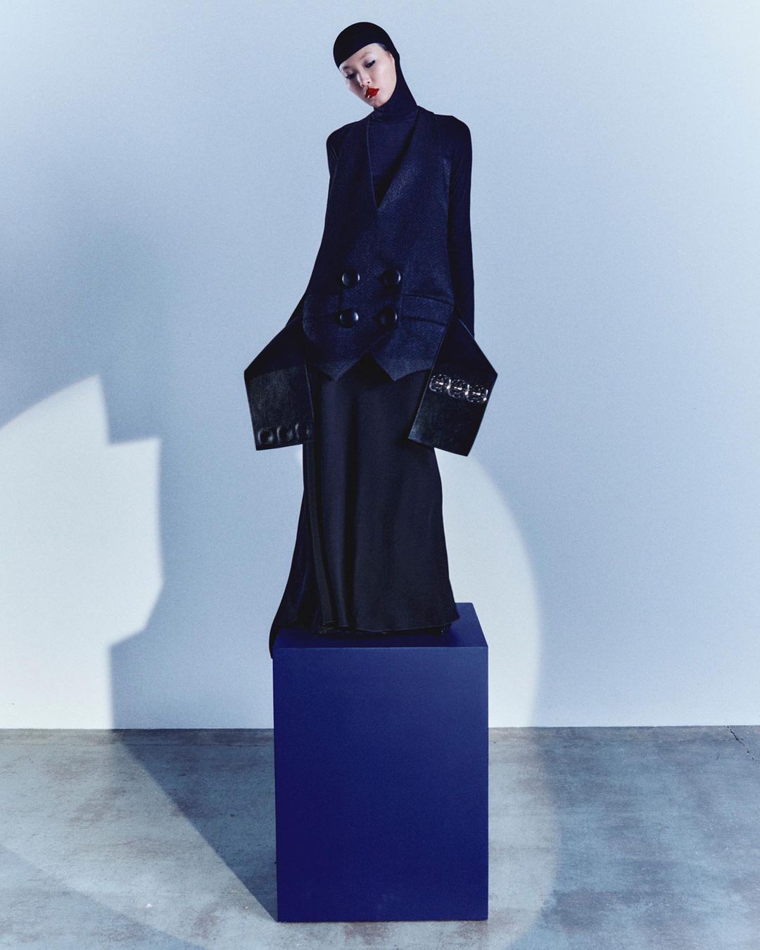 Fashion shot of a woman in a black jacket and long skirt standing on a blue cube against a gradient blue studio backdrop.