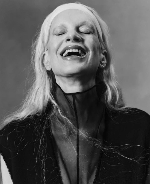 Black and white portrait of a smiling adult female with a headband, closed eyes, nose piercing, and sheer black top against a neutral background.