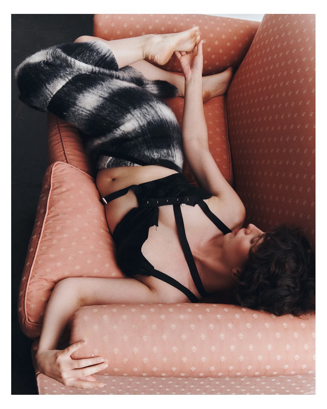 Woman lounges in pink armchair wearing a black and white furry garment and lingerie. Relaxed pose, soft lighting, indoors.