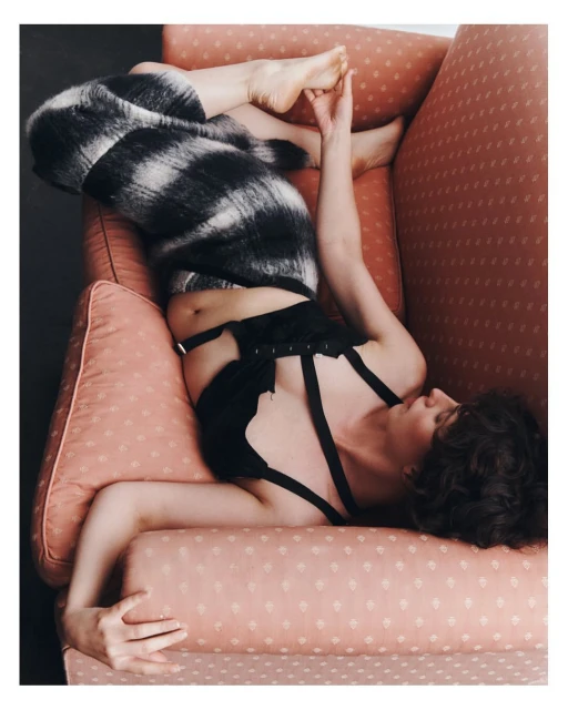 Woman lounges in pink armchair wearing a black and white furry garment and lingerie. Relaxed pose, soft lighting, indoors.
