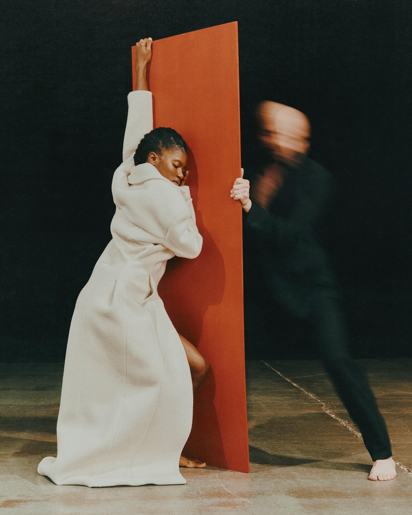 Two figures lean against a bright red panel indoors: A Black female adult in long white coat with bare feet and a blurry male figure.