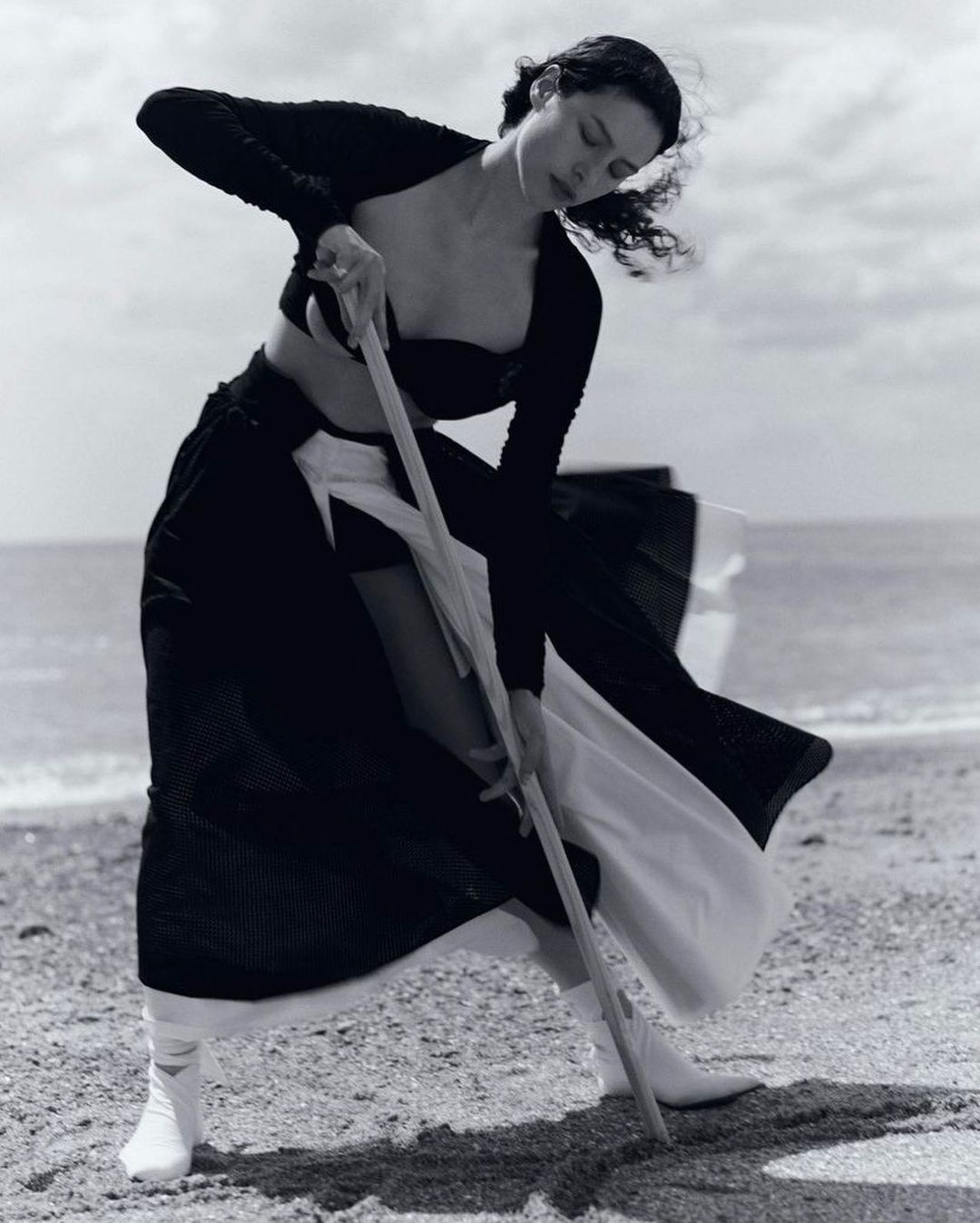 A woman on a beach holds a long pole, digging into the sand. The black and white photo features the sea and cloudy sky in the background.