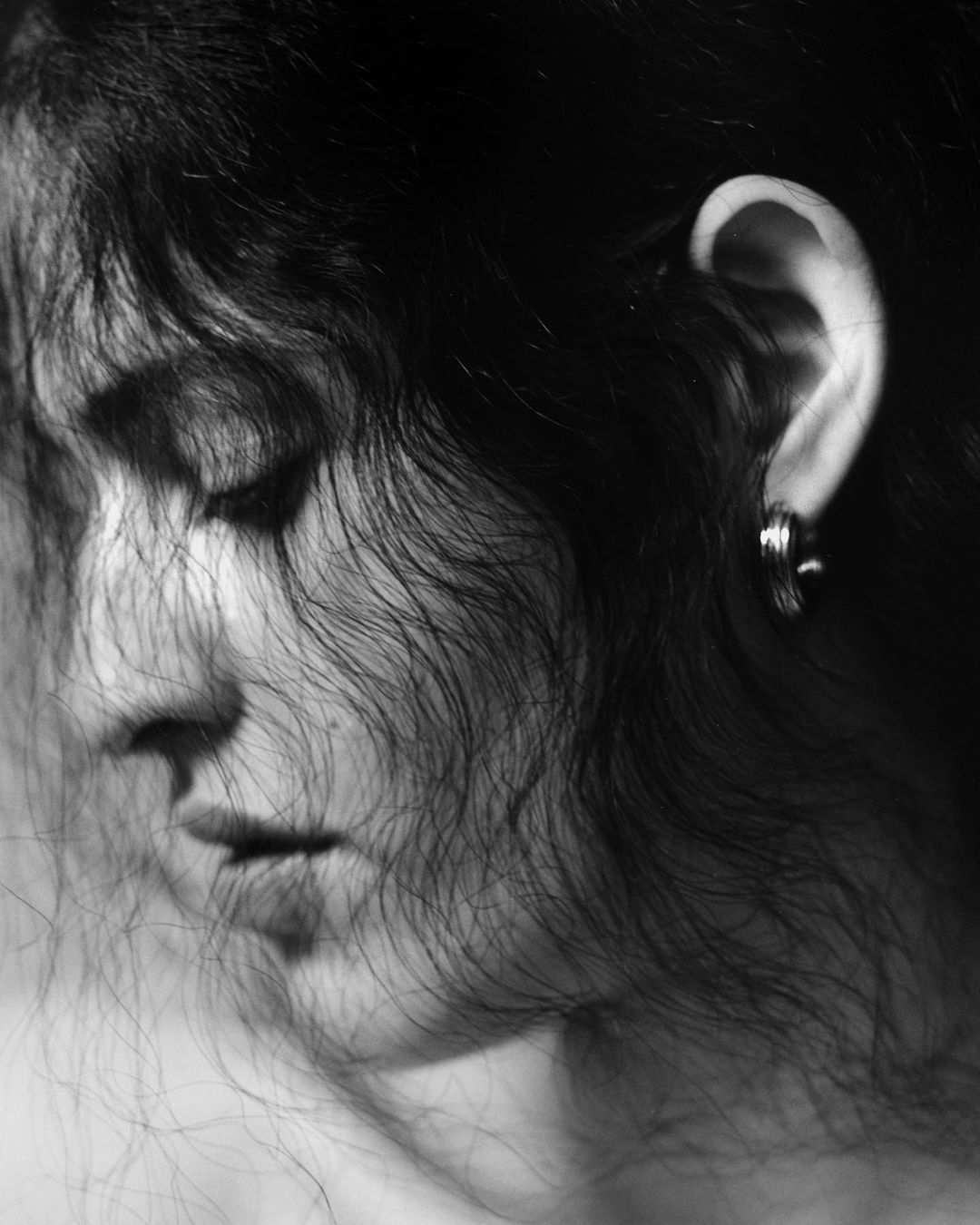 Black and white portrait of a serene adult woman with dark curly hair and a hoop earring, softly lit with a dark background.