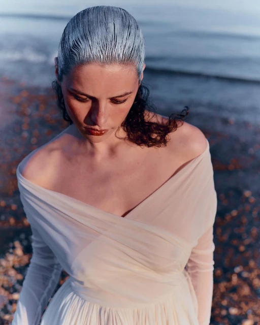 Young woman with silver hair and an off-shoulder dress stands on a pebbled beach with ocean water in the background.