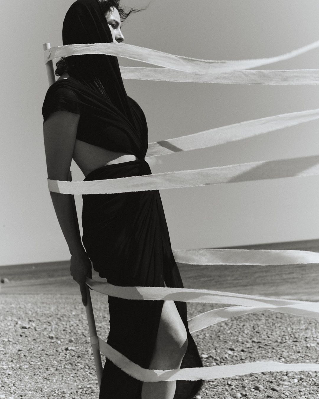 Monochrome shot of a woman wrapped in fabric standing on a beach, featuring ribbons, a shore, and a serene sky, creating a dramatic scene.
