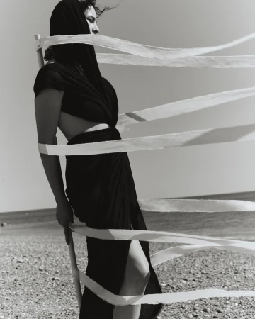 Monochrome shot of a woman wrapped in fabric standing on a beach, featuring ribbons, a shore, and a serene sky, creating a dramatic scene.