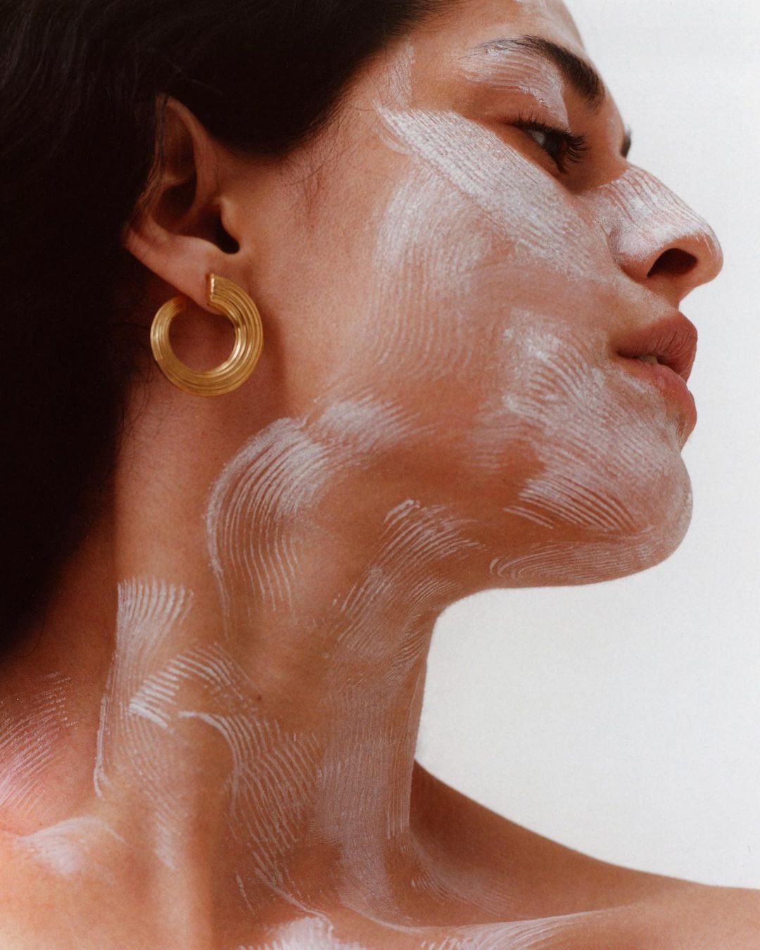 Close-up portrait of a woman with cream on her face, wearing gold hoop earrings, posing in front of a white background.