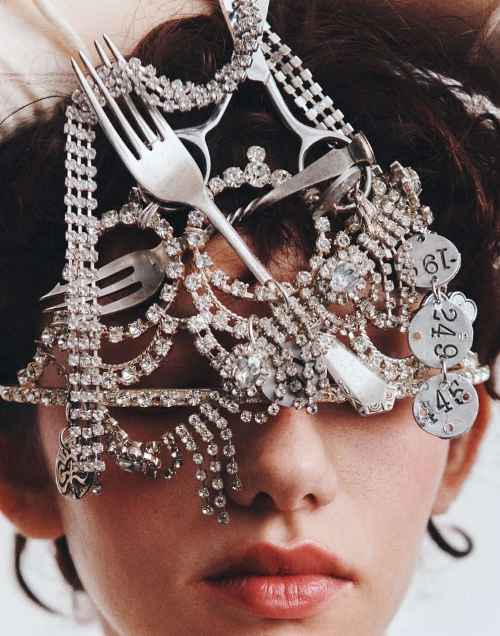 Woman with elaborate headwear featuring forks, jewelry, and metal decorations, creating an eye-obscuring accessory against a bright background.