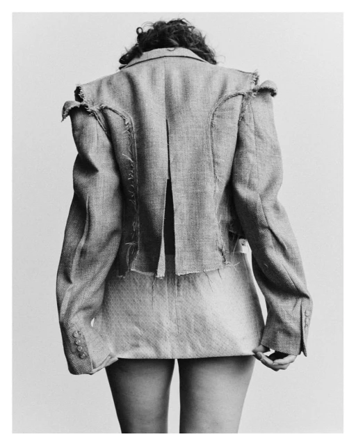 Back view of an adult female wearing a light-colored tweed jacket and skirt in a studio setting with a soft, neutral background.