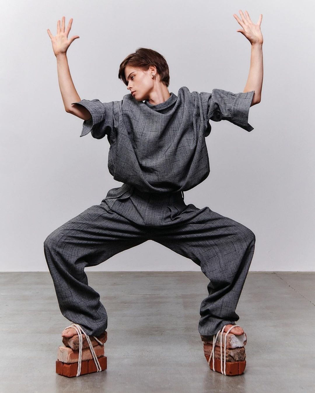 Fashion-forward individual strikes a pose in matching gray ensemble, unconventional brick footwear, set against a neutral backdrop. 