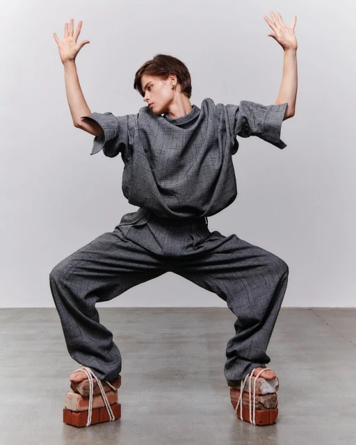 Fashion-forward individual strikes a pose in matching gray ensemble, unconventional brick footwear, set against a neutral backdrop. 