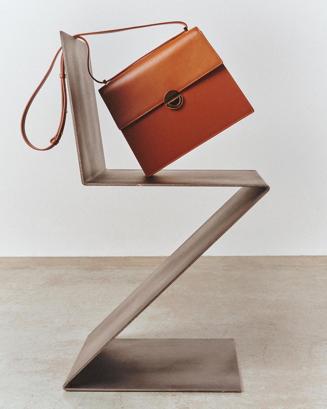 Studio shot of an orange leather satchel handbag with strap on a modern zig-zag chair against a neutral backdrop and smooth concrete floor.