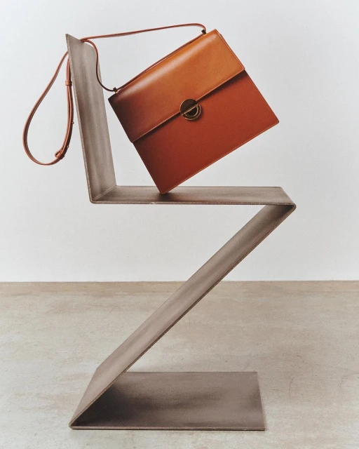 Studio shot of an orange leather satchel handbag with strap on a modern zig-zag chair against a neutral backdrop and smooth concrete floor.