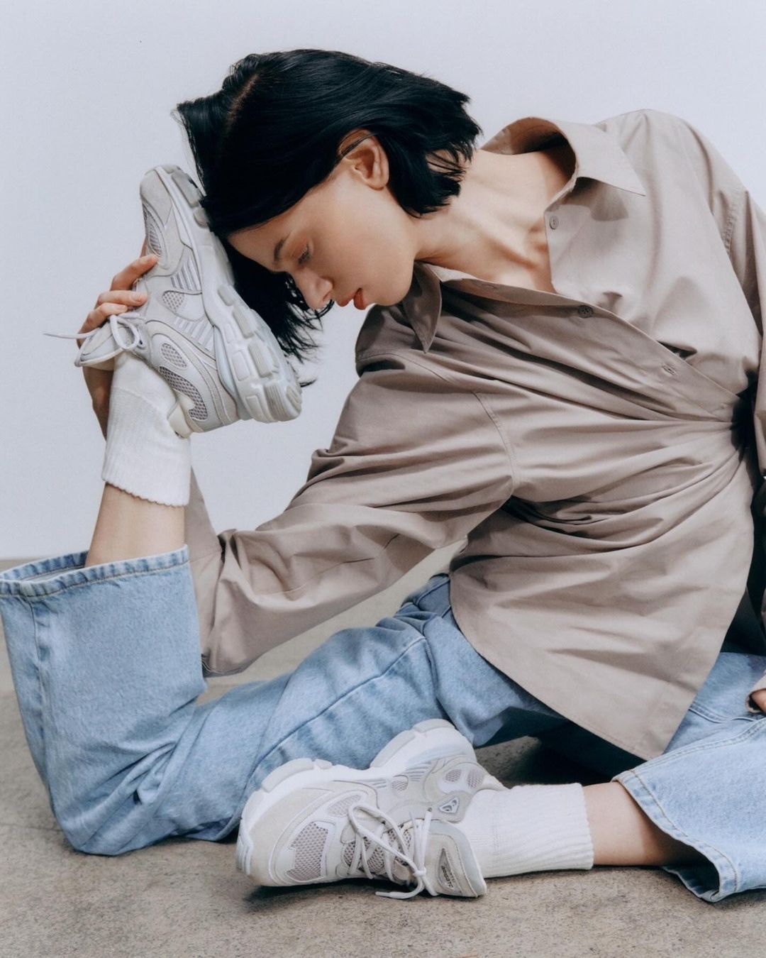 A person in a beige shirt and blue jeans sits on the floor, leg raised to head with the foot resting on their forehead, and sneakers on.