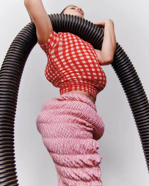 A woman poses in a red and white checkered top and pink and white checkered pants, with a large black corrugated tube around her neck.