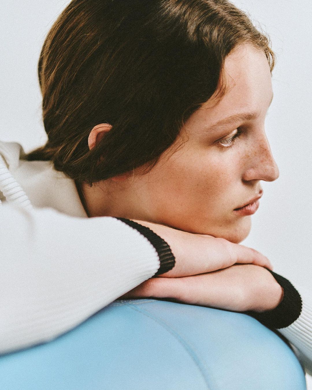 Close-up portrait of a young fair-skinned woman with dark hair, resting her arms on a light blue exercise ball in a minimalist setting.