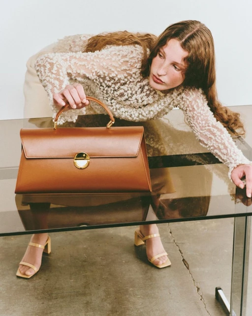 Woman presents a brown leather top-handle bag, wearing a lace top and light yellow high heels, in a bright studio setting.