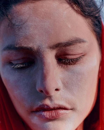 Close-up portrait of a woman with powdered skin and closed eyes, framed by a vibrant red head covering, photographed by Lara Angelil.