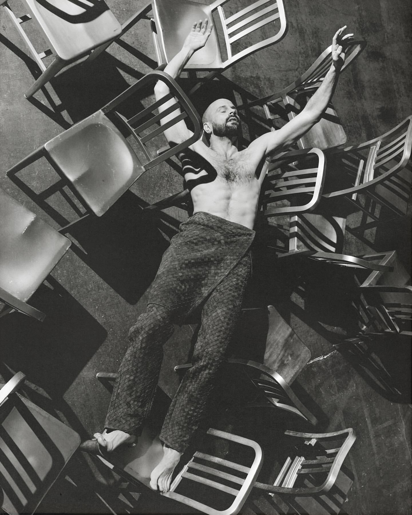 Monochrome portrait of a shirtless man lying on a pile of chairs with arms raised in an indoor setting with a dark background.